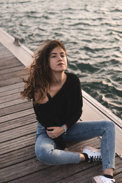  mladá žena, zatímco sedí na dřevěném molu blízko vody při pohledu na fotoaparát - Fotografie, Obrázek