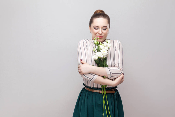 Retrato de atractiva mujer joven placer romántico en camisa a rayas y falda verde abrazando ramo de flores blancas sobre fondo gris
  - Foto, imagen