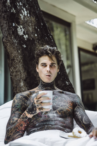 young handsome tattooed man having breakfast in bed in the open air outdoors in the garden, donuts and coffee for breakfast. - Foto, afbeelding