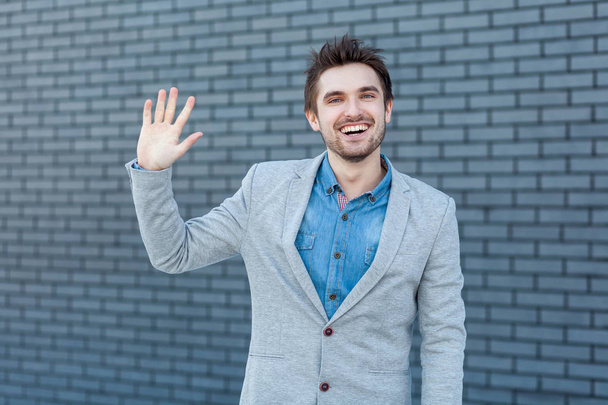 Portrait of happy handsome bearded man in casual style standing and looking at camera with greeting gesture and toothy smiling on brick wall background. - Zdjęcie, obraz
