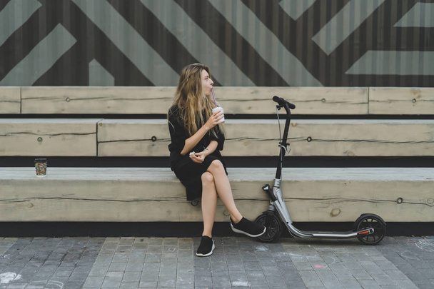 Young woman with long hairs on electric scooter. The girl on the electric scooter drinks coffee. - Φωτογραφία, εικόνα