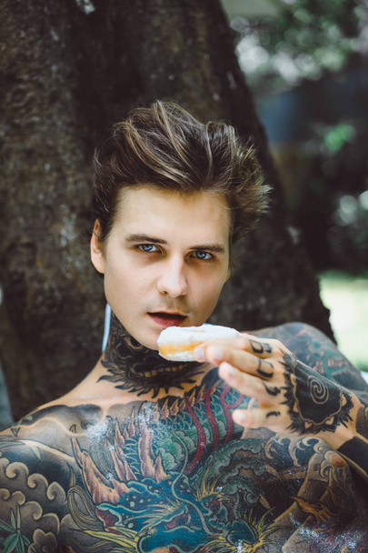 Молодой красивый татуированный мужчина завтракает в постели на открытом воздухе в саду, пончики и кофе на завтрак
. - Фото, изображение