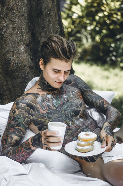 young handsome tattooed man having breakfast in bed in the open air outdoors in the garden, donuts and coffee for breakfast. - Valokuva, kuva