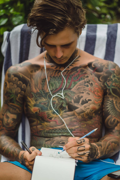a young man in tattoos wearing headphones listens to music and draws a pencil in a notebook. Online training. - Foto, immagini