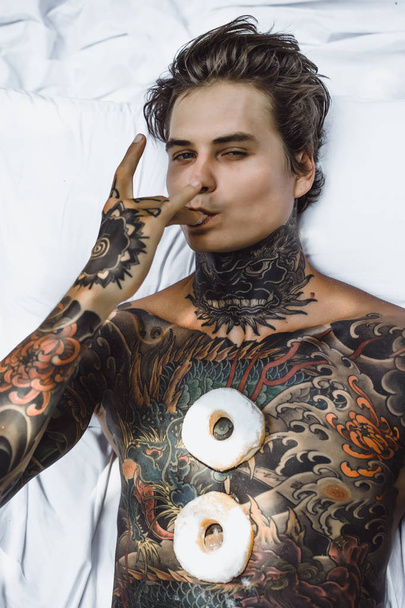 young handsome tattooed man having breakfast in bed in the open air outdoors in the garden, donuts and coffee for breakfast. - Фото, зображення