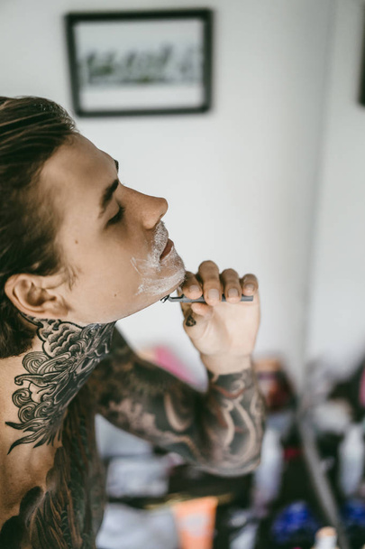 a young handsome tattooed man in the morning shaves in front of a mirror, a razor in his hands, a shave. - Fotó, kép