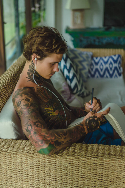 a young man in tattoos wearing headphones listens to music and draws a pencil in a notebook. Online training. - Foto, Imagen