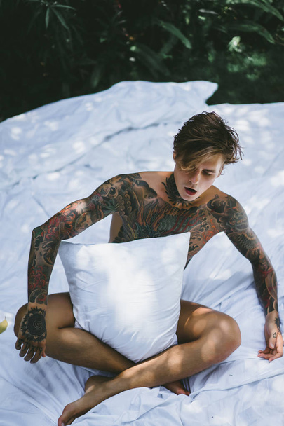 a young handsome tattooed man in the morning in bed tosses up the pillows. bed in the garden - Photo, image