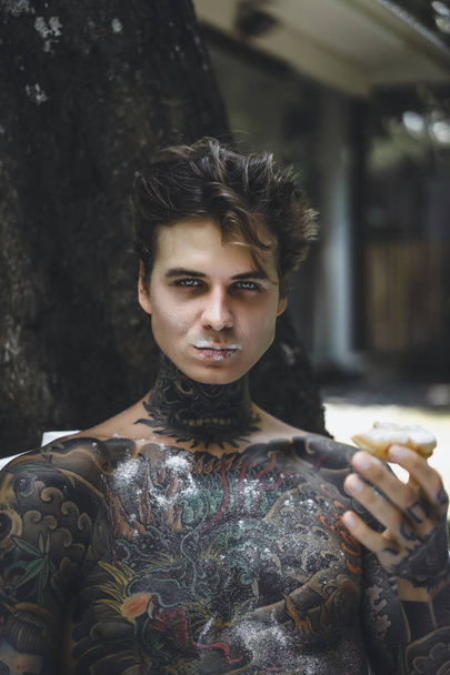 young handsome tattooed man having breakfast in bed in the open air outdoors in the garden, donuts and coffee for breakfast. - 写真・画像
