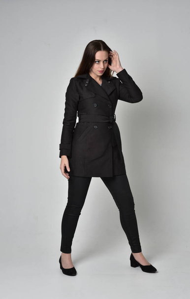 灰色のスタジオの背景に立ちポーズ長く黒いコートを着ているブルネットの少女の完全な長さの肖像画. - 写真・画像