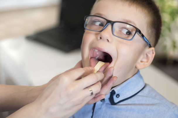 Ένα αγόρι με το στόμα ανοιχτό κατά τη διάρκεια μιας ιατρικής εξέτασης. Ο γιατρός ελέγχει το λαιμό και τις αμυγδαλές του ασθενή με μια ξύλινη σπάτουλα. - Φωτογραφία, εικόνα