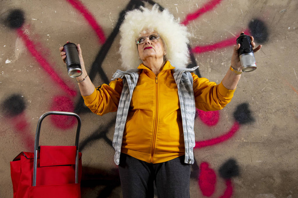 cool rebel grandmother painting graffiti against an urban wall - Photo, Image