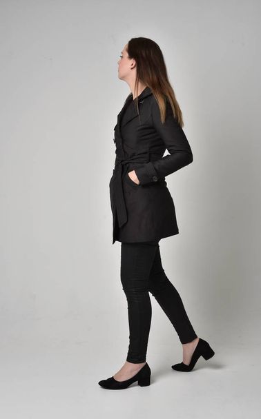 портрет брюнетки в длинном черном пальто, стоящей в позе спиной к камере на сером фоне студии
. - Фото, изображение