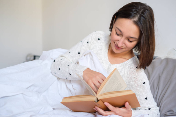 Молода дівчина читає книгу, що лежить у ліжку
 - Фото, зображення