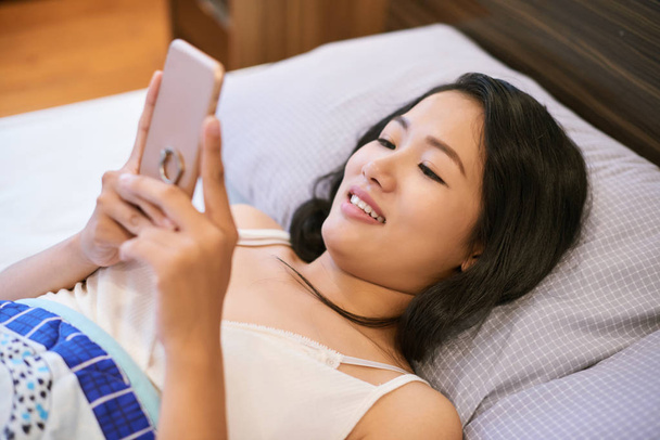 From above of young Asian woman surfing phone while lying in bed in morning time - Fotoğraf, Görsel