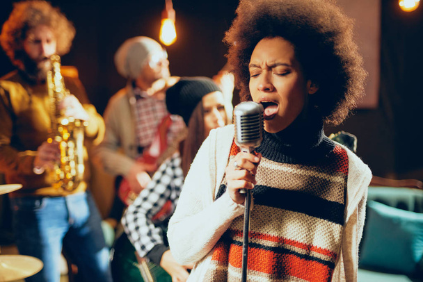 Detailní záběr Smíšené rasy žena zpívá. V pozadí kapela hrající nástroje. Domácí studio interiér. - Fotografie, Obrázek