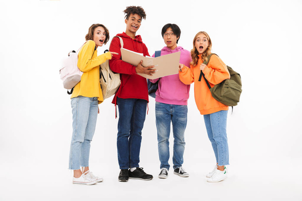 Group of cheerful teenagers isolated over white background, carrying backpacks, looking at an open folder - Фото, изображение