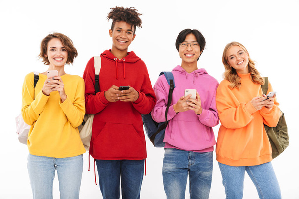 Group of cheerful teenagers isolated over white background, carrying backpacks, using mobile phones - Foto, imagen