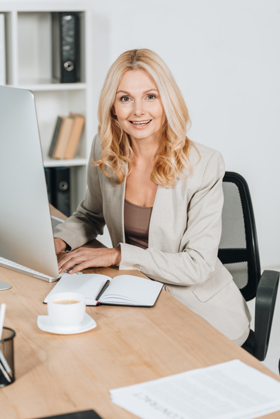femme d'affaires mature gaie utilisant un ordinateur de bureau et souriant à la caméra dans le bureau
 - Photo, image