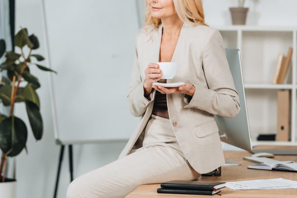 обрезанный снимок улыбающейся деловой женщины, держащей чашку кофе и сидящей на столе в офисе
 - Фото, изображение
