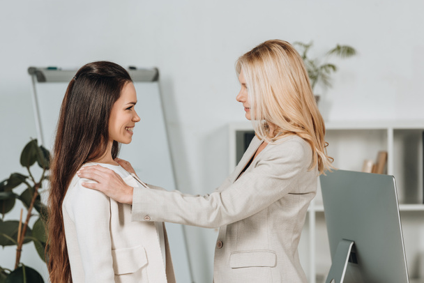 side view of mature businesswoman supporting smiling young female colleague in office - Photo, image