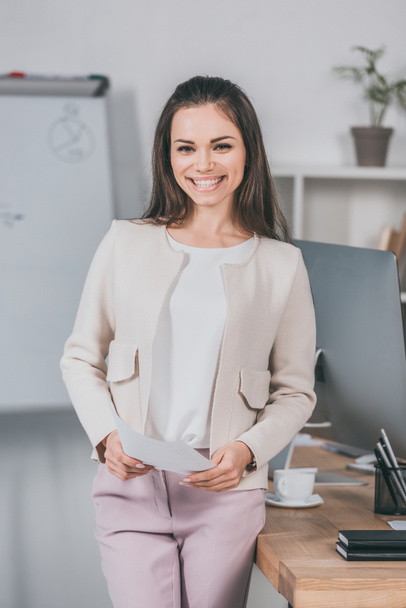 beautiful happy young businesswoman holding papers and smiling at camera in office - Valokuva, kuva