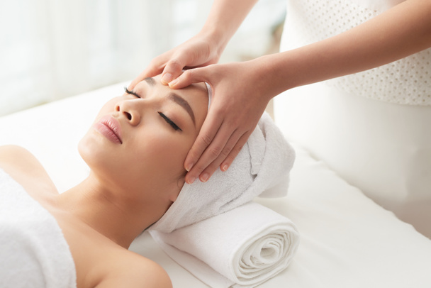 terapeuta anónima frotando la frente de atractiva cliente femenina durante la sesión de masaje en el salón de spa
 - Foto, imagen
