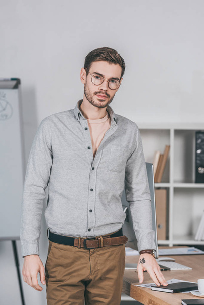 handsome professional young businessman in eyeglasses standing and looking at camera in office - Foto, afbeelding
