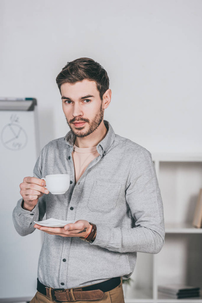 handsome young businessman holding cup of coffee and looking at camera in office  - Foto, immagini