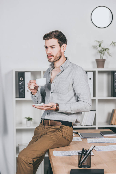 young businessman holding cup of coffee and sitting on desk in office - Fotó, kép