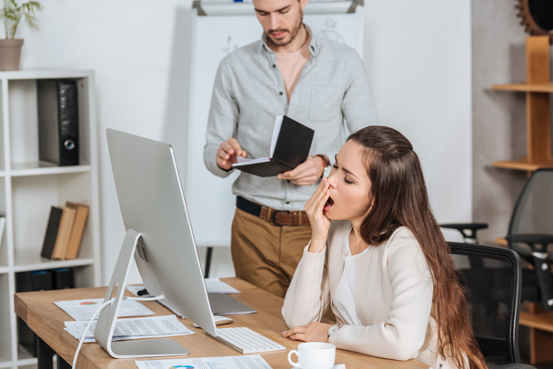 business mentor holding notebook and young female colleague yawning at workplace - Photo, Image