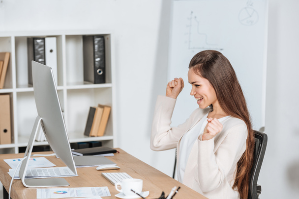 Seitenansicht einer glücklichen jungen Geschäftsfrau, die im Büro die Fäuste schüttelt und auf den Desktop-Computer blickt - Foto, Bild