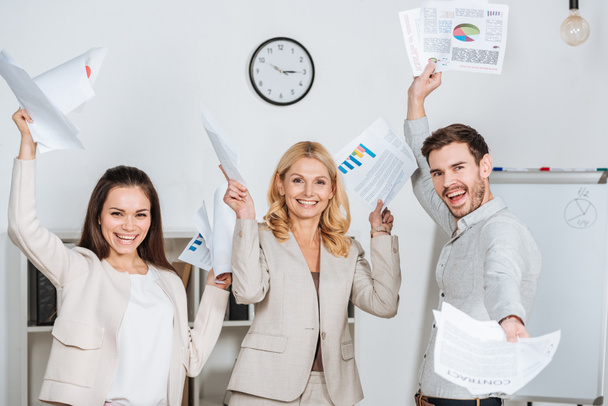 веселые профессиональные деловые коллеги, держащие бумаги и улыбающиеся перед камерой в офисе
 - Фото, изображение