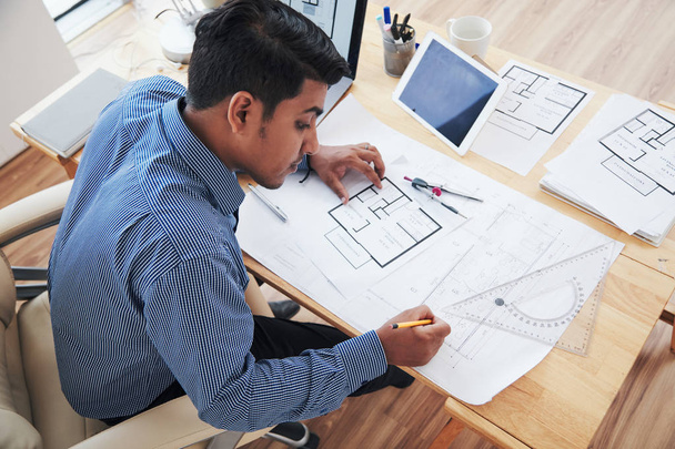 Felülről felnőtt indiai építész dolgozik blueprint, miközben ül íróasztal hivatalban - Fotó, kép