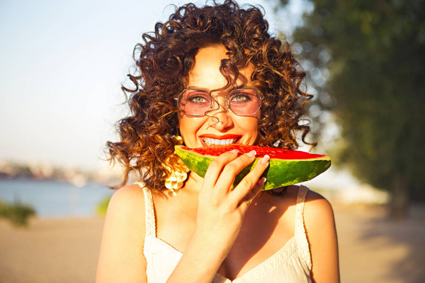 Kauneus hymyilevä kihara nainen on yllään vaaleanpunainen aurinkolasit ja syö vesimelonia rannalla
 - Valokuva, kuva