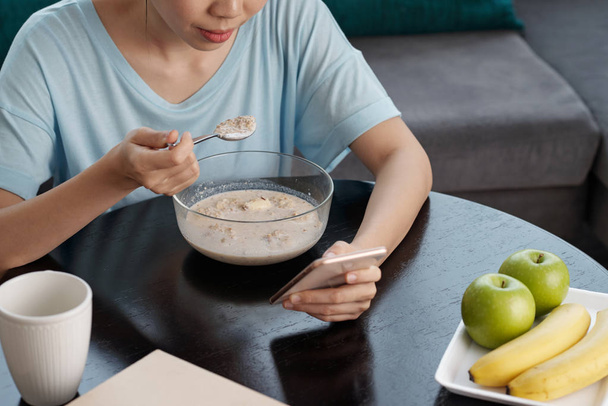 Shůry záběr anonymní lady procházení smartphone a jíst müsli zatímco sedí u stolu v obývacím pokoji - Fotografie, Obrázek