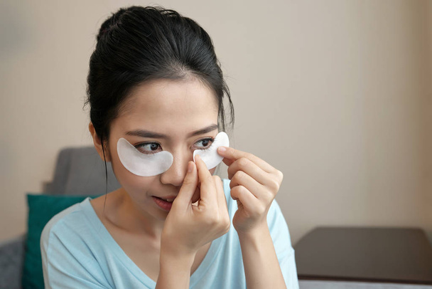 Attractive Asian woman applying under-eye patches while sitting in stylish living room - Фото, изображение