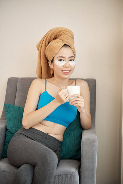 Charming Asian woman with under-eye patches smiling and looking at camera while enjoying hot beverage in comfortable armchair - Photo, Image