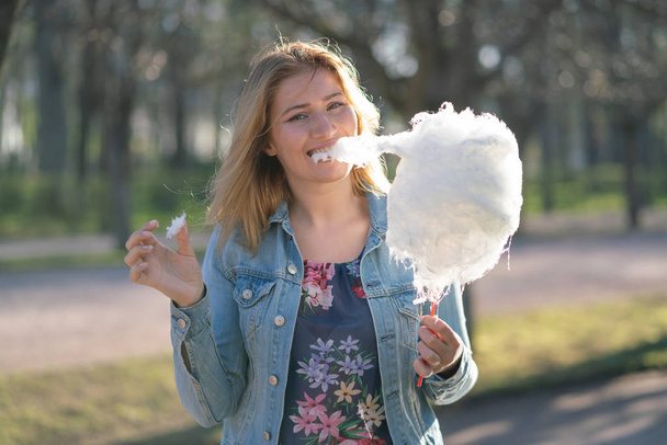 charming caucasian girl walks in the spring Park in a denim jacket and eats pink cotton candy, smiles at the morning sunlight - Photo, Image