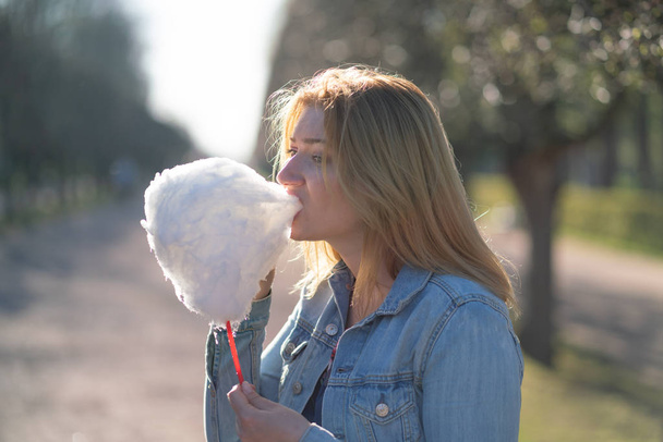 charming caucasian girl walks in the spring Park in a denim jacket and eats pink cotton candy, smiles at the morning sunlight - Foto, imagen