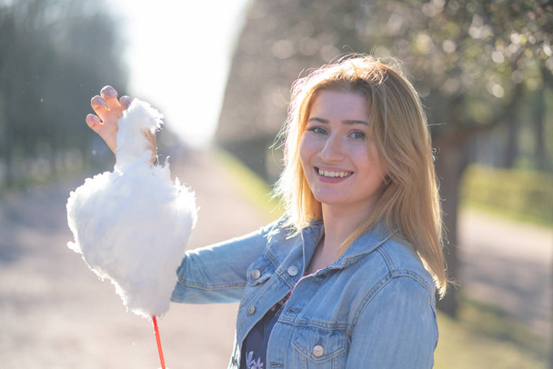 charming caucasian girl walks in the spring Park in a denim jacket and eats pink cotton candy, smiles at the morning sunlight - Fotoğraf, Görsel