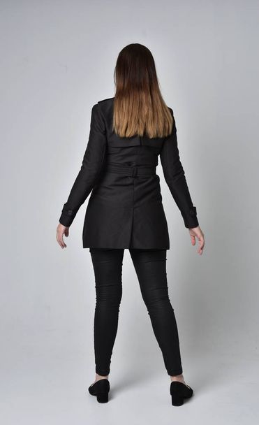 full length portrait of a brunette girl wearing long black coat, standing pose with back to the camera on grey studio background. - Фото, зображення