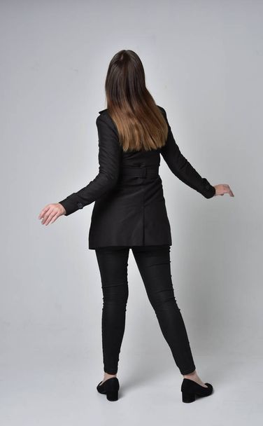 full length portrait of a brunette girl wearing long black coat, standing pose with back to the camera on grey studio background. - Фото, зображення