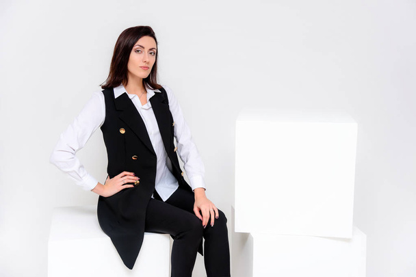 Concept portrait of a girl sitting on a white background in a business suit in different poses with different emotions. It in the middle of the frame, next to the white cubes, next is another cube. - Foto, imagen