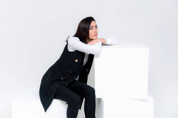 Concept portrait of a girl sitting on a white background in a business suit in different poses with different emotions. It in the middle of the frame, next to the white cubes, next is another cube. - Foto, Imagen