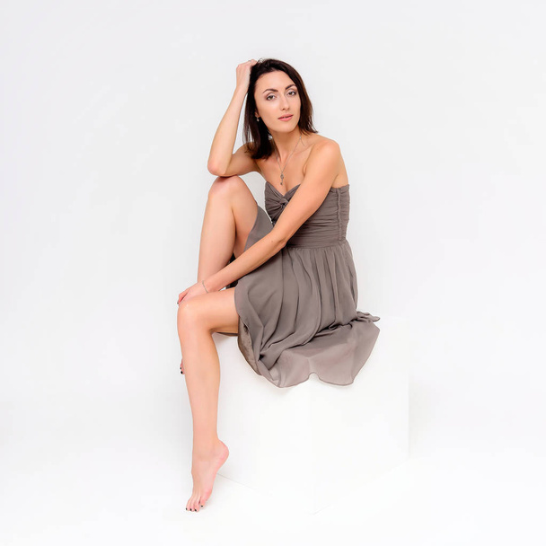 Concept portrait of a brunette girl, sitting on a white background in a beautiful brown dress on a white cube in studio. She sits right in front of the camera in various poses with different emotions. - Foto, afbeelding