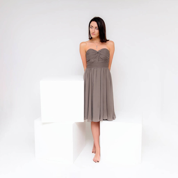 Concept portrait of a brunette girl, standing on a white background in a beautiful brown dress near the white cube. She sits right in front of the camera in various poses with different emotions. - Fotoğraf, Görsel