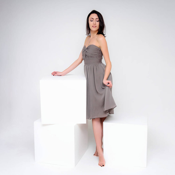 Concept portrait of a brunette girl, standing on a white background in a beautiful brown dress near the white cube. She sits right in front of the camera in various poses with different emotions. - Foto, Bild