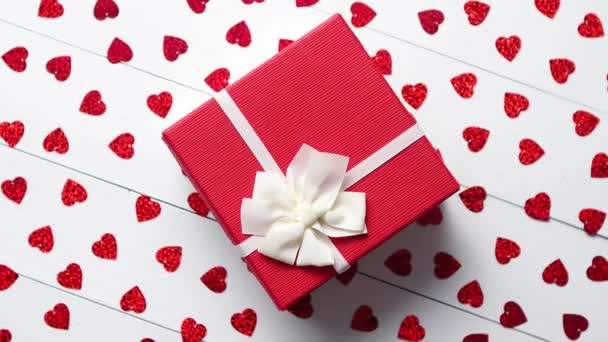 Valentýn dekorace složení. Zabalený dárek kladen na červené flitry na bílý dřevěný stůl ve tvaru srdce. Romantické pozadí. Plochá ležel, horní pohled. - Záběry, video