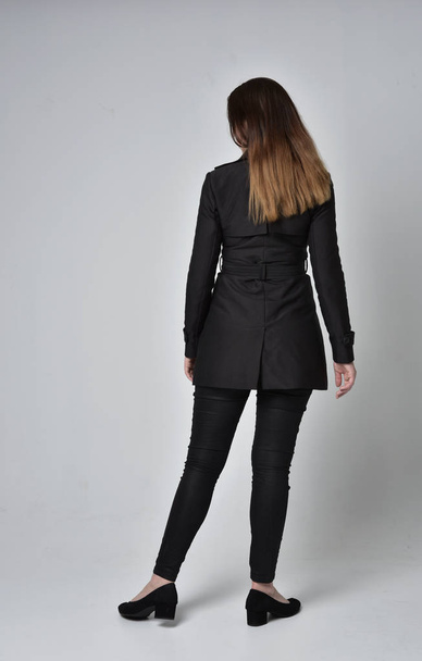 full length portrait of a brunette girl wearing long black coat, standing pose with back to the camera on grey studio background. - Fotó, kép